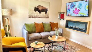 A living room with a sofa, two side tables, a coffee table, a lamp, a colorful rug, a framed picture of a bison, and a wall-mounted TV displaying travel destinations. - Modern Downtown Suite - Walking Distance to Restaurants, Shops, & Grocery Stores (Golden)