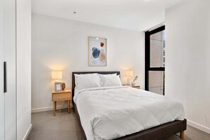 A bedroom with a bed, two bedside tables with lamps, a framed artwork above the bed, and a window with a view of a brick building.