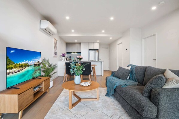 A modern living room with a flat-screen TV, a grey sofa, a wooden coffee table, and a dining area with a kitchen in the background.
