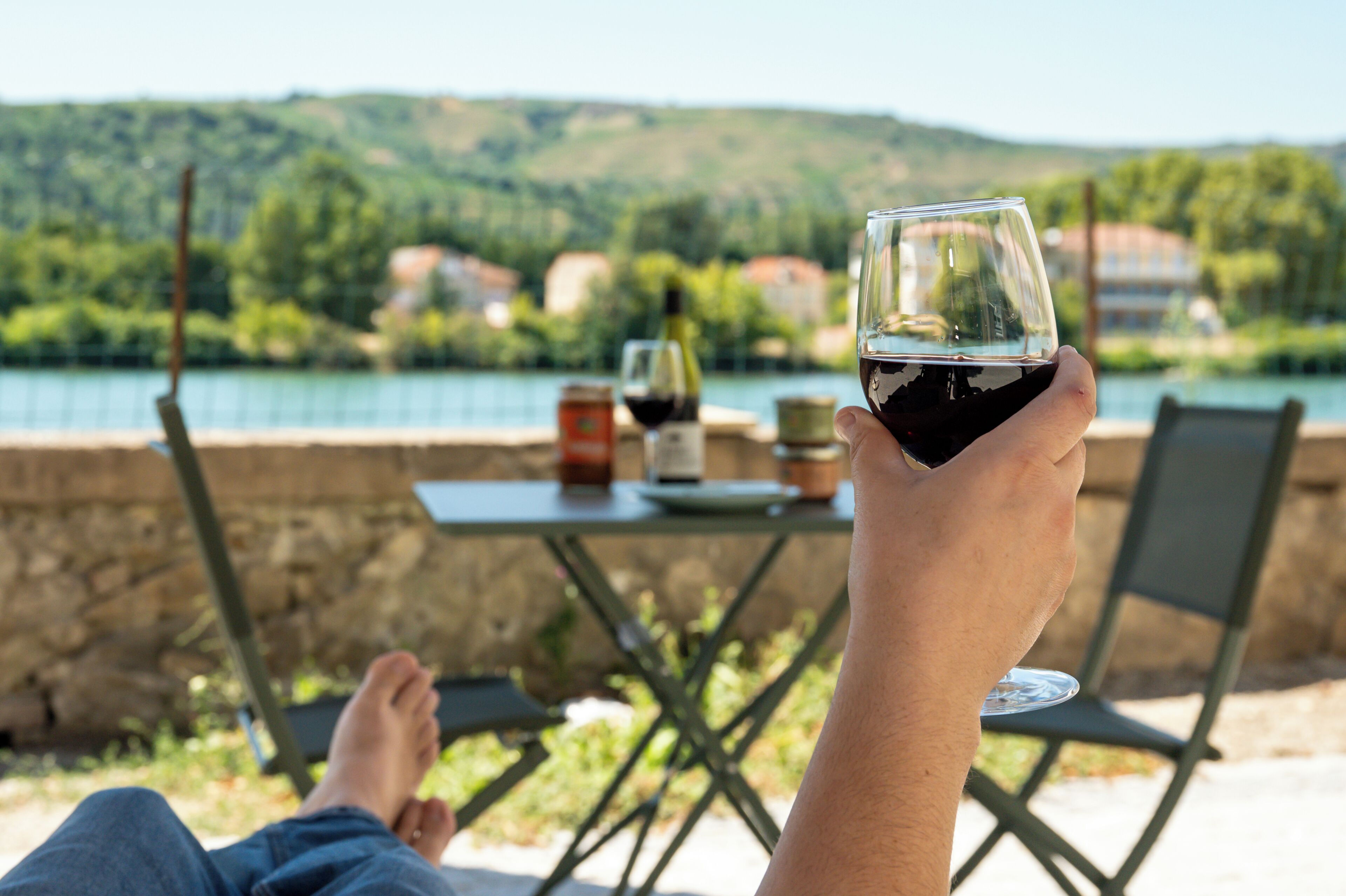 Une personne se détend à une table au bord d’un lac, un verre de vin rouge à la main, et profite d’une vue panoramique sur les maisons et les collines environnantes.