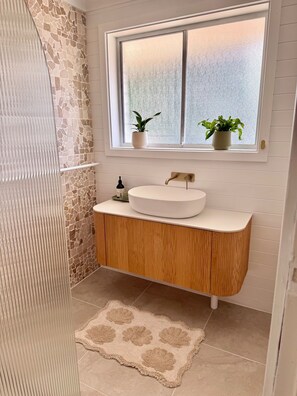 Un baño con pared de mosaico de piedra, un lavamanos blanco con tocador de madera, una ventana con dos plantas en macetas y una alfombra color beige.