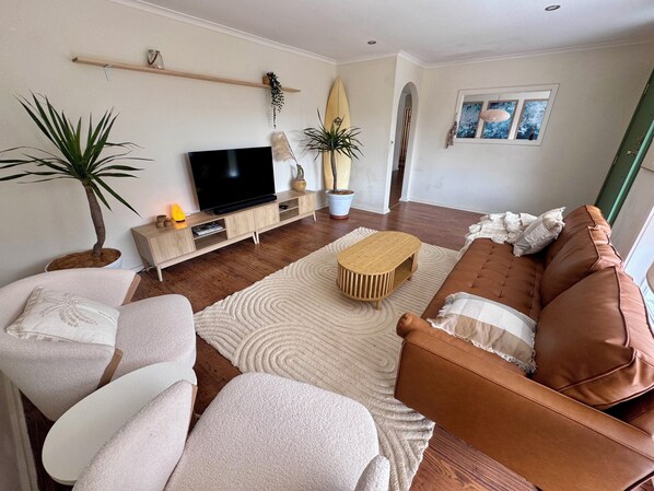 Un salón con un sofá de cuero marrón, sillones blancos, un televisor de pantalla plana, estanterías de madera, una planta en maceta y una tabla de surf.