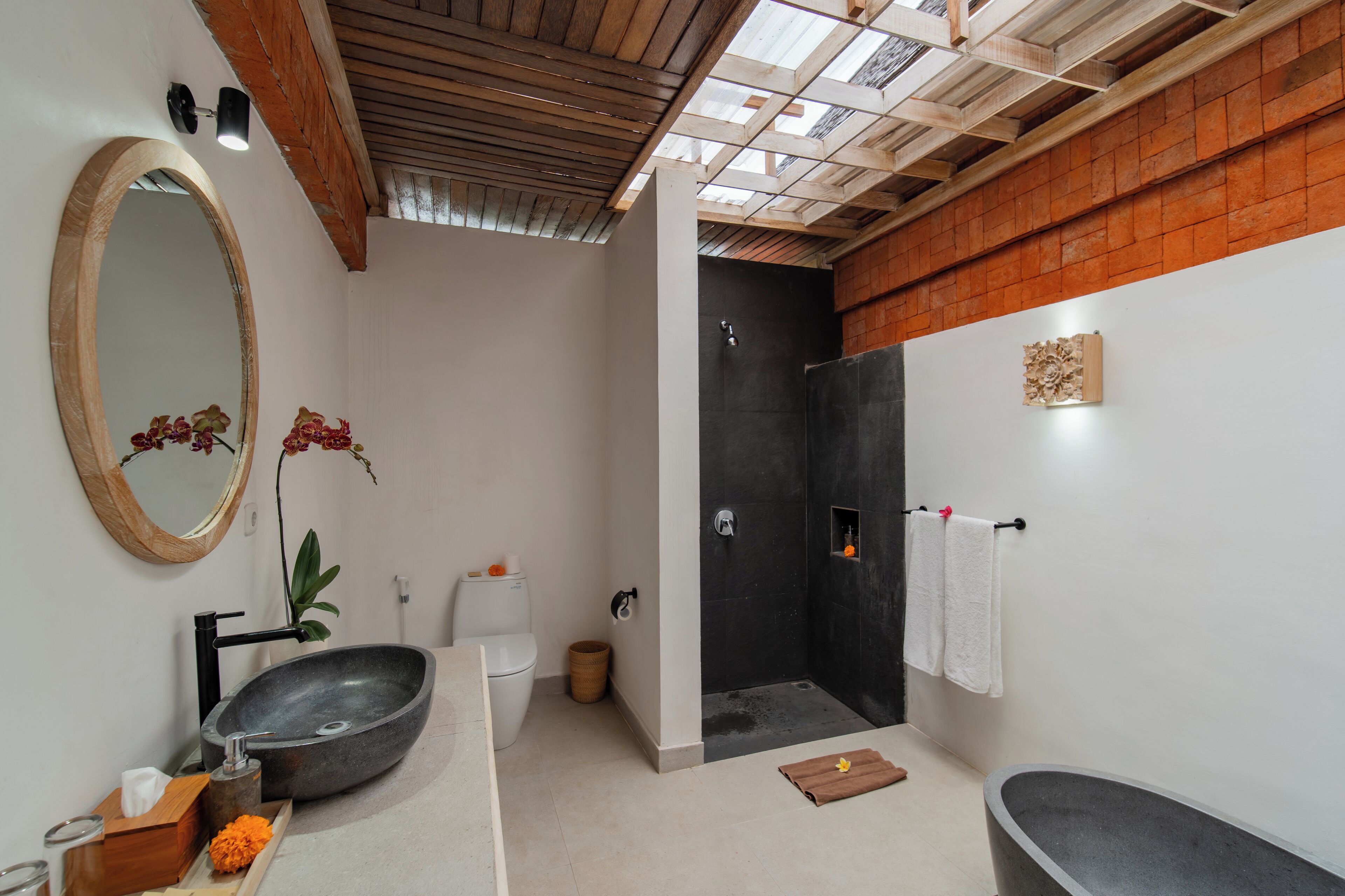 A modern bathroom with a large oval mirror, a stone sink, a freestanding bathtub, and a walk-in shower.