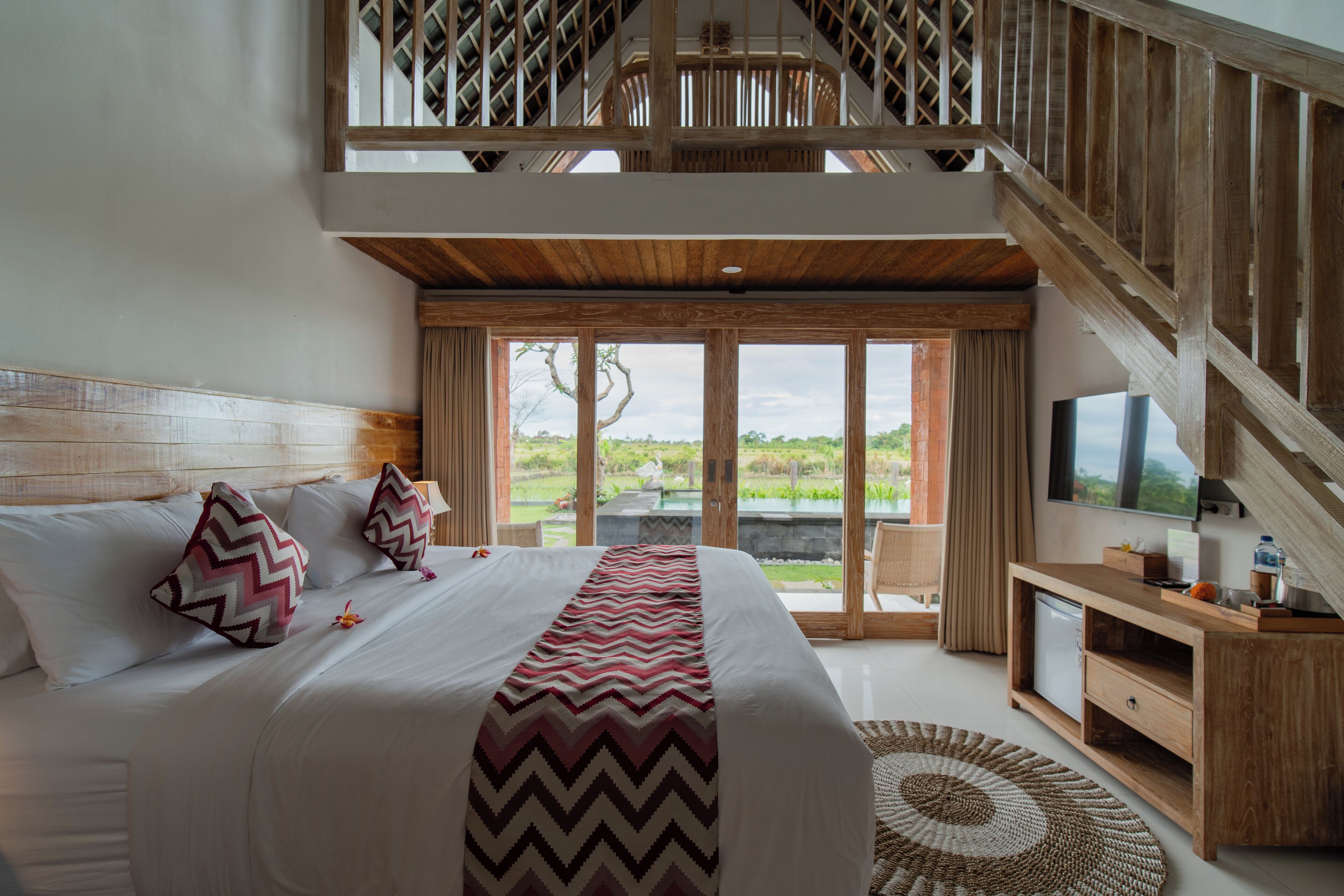 Une chambre spacieuse avec un grand lit, un bureau en bois, une télévision et un balcon offrant une vue sur la verdure.