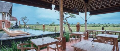 An outdoor patio with wooden tables and chairs, a pool, and a view of a field.