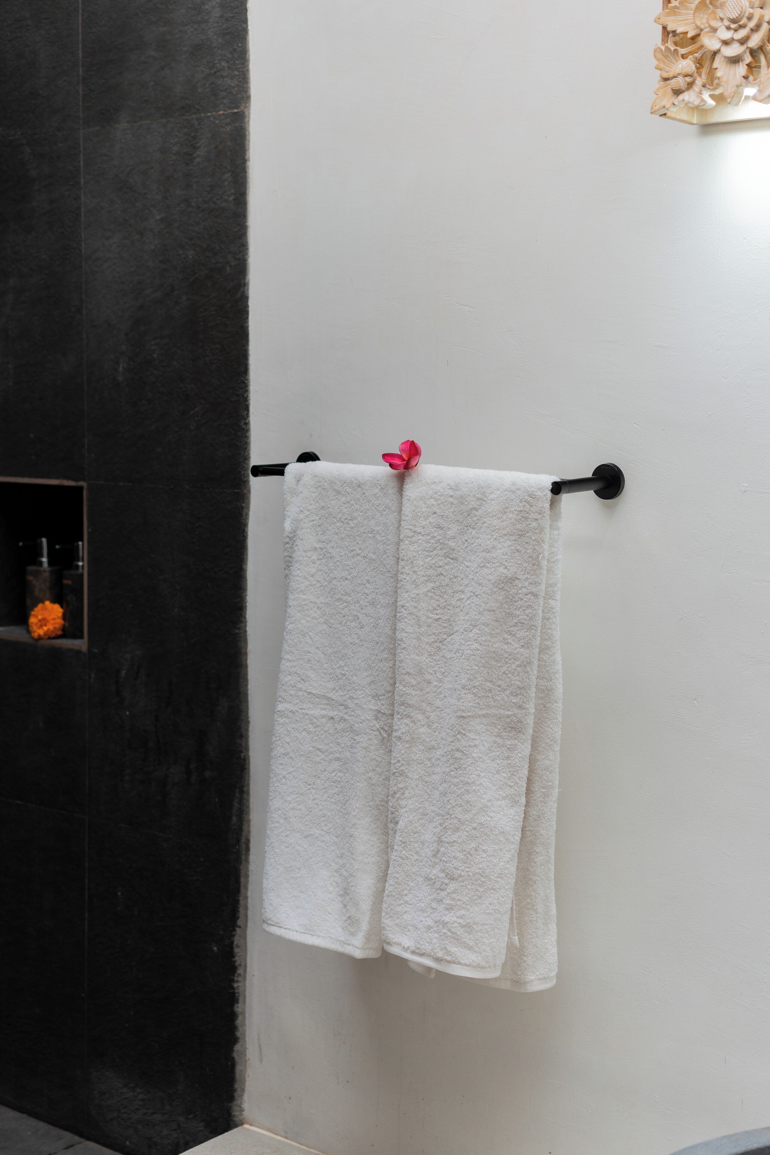 A bathroom with a black wall, a white towel hanging on a black towel rack, and a small shelf with a glass container and orange flowers.