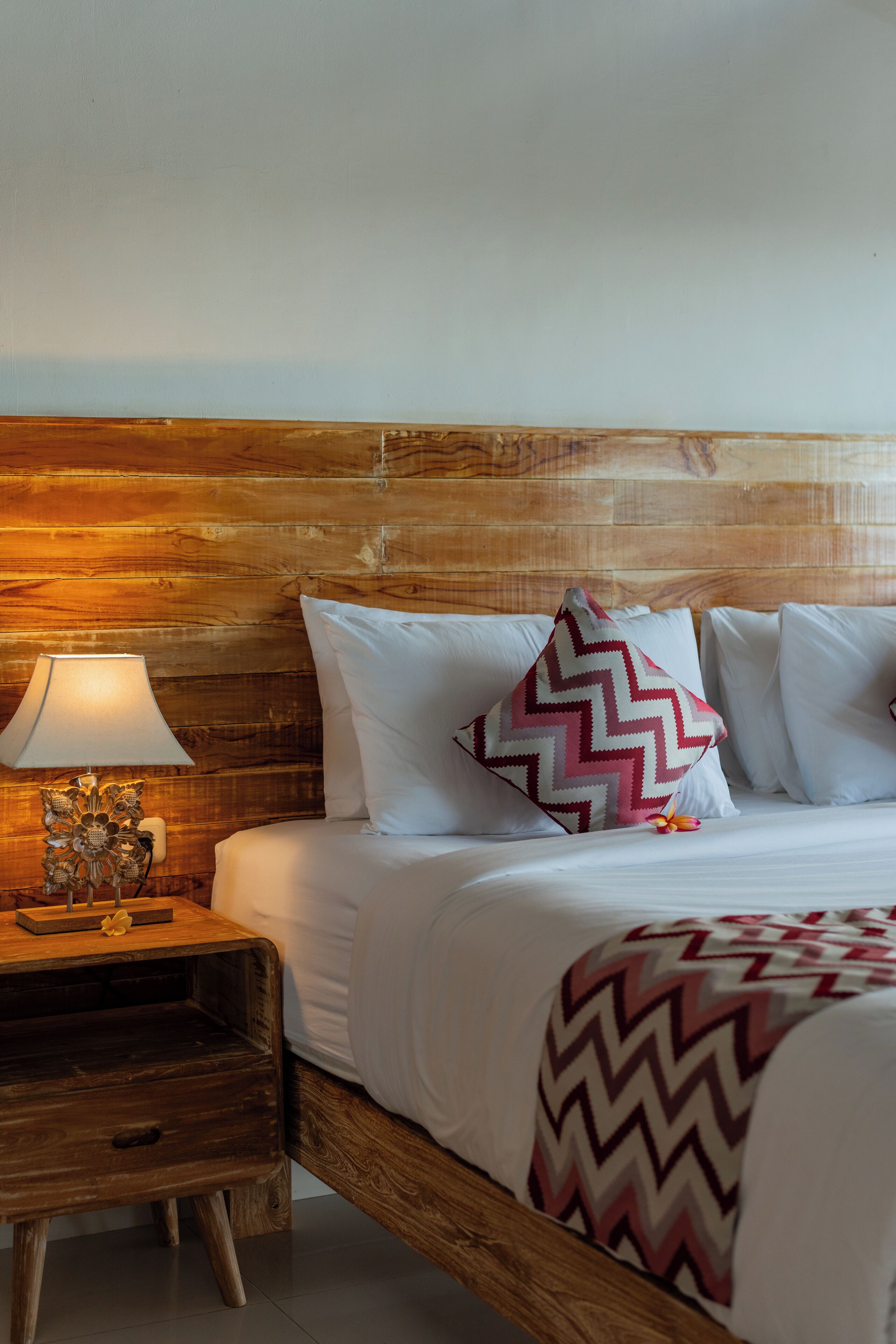 Una cama bien hecha con sábanas blancas y un edredón estampado, una mesita de noche de madera con una lámpara y un cabecero de madera.