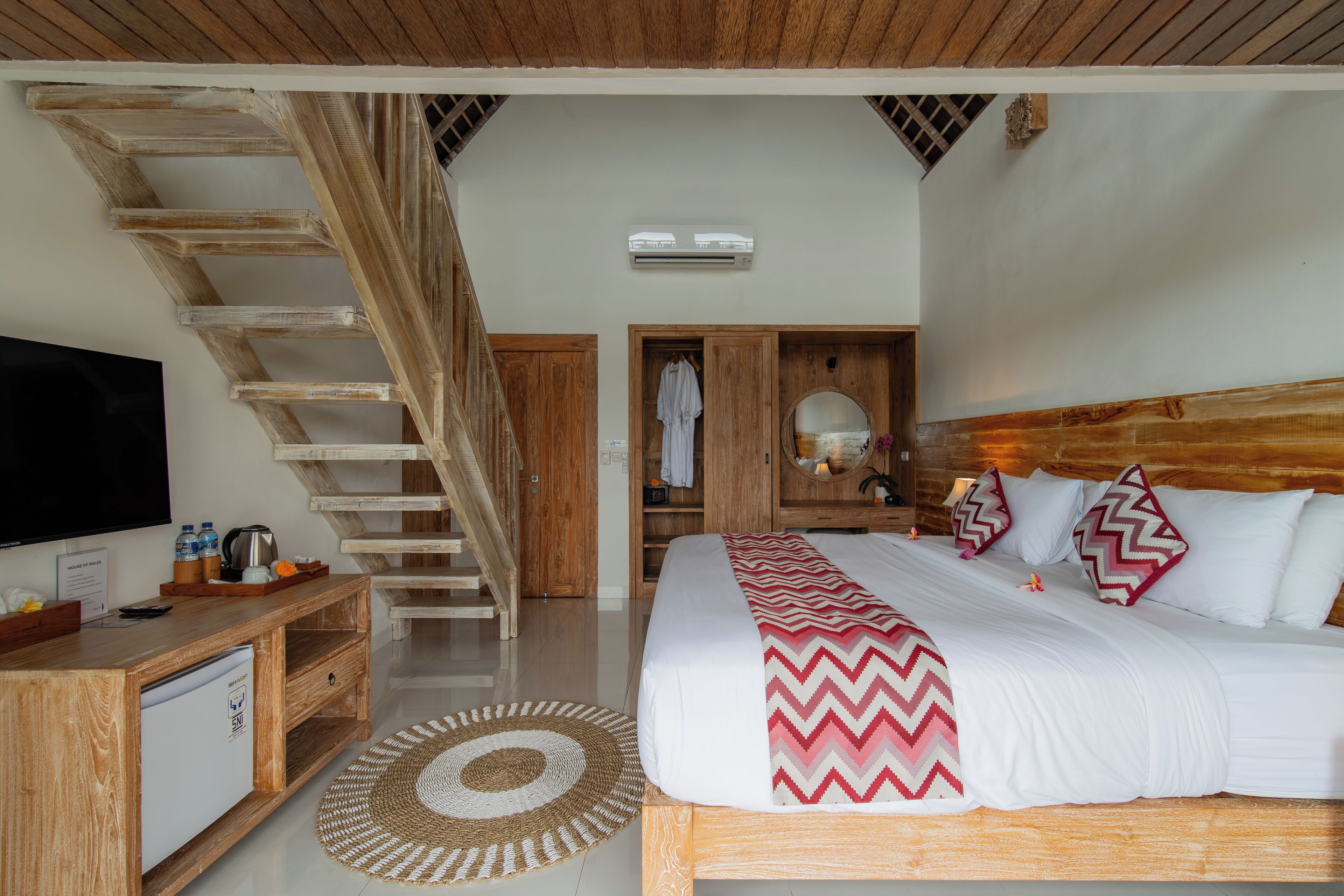 A bedroom with a wooden staircase, a bed with a patterned blanket, a bedside table with a kettle, and a flat-screen TV.