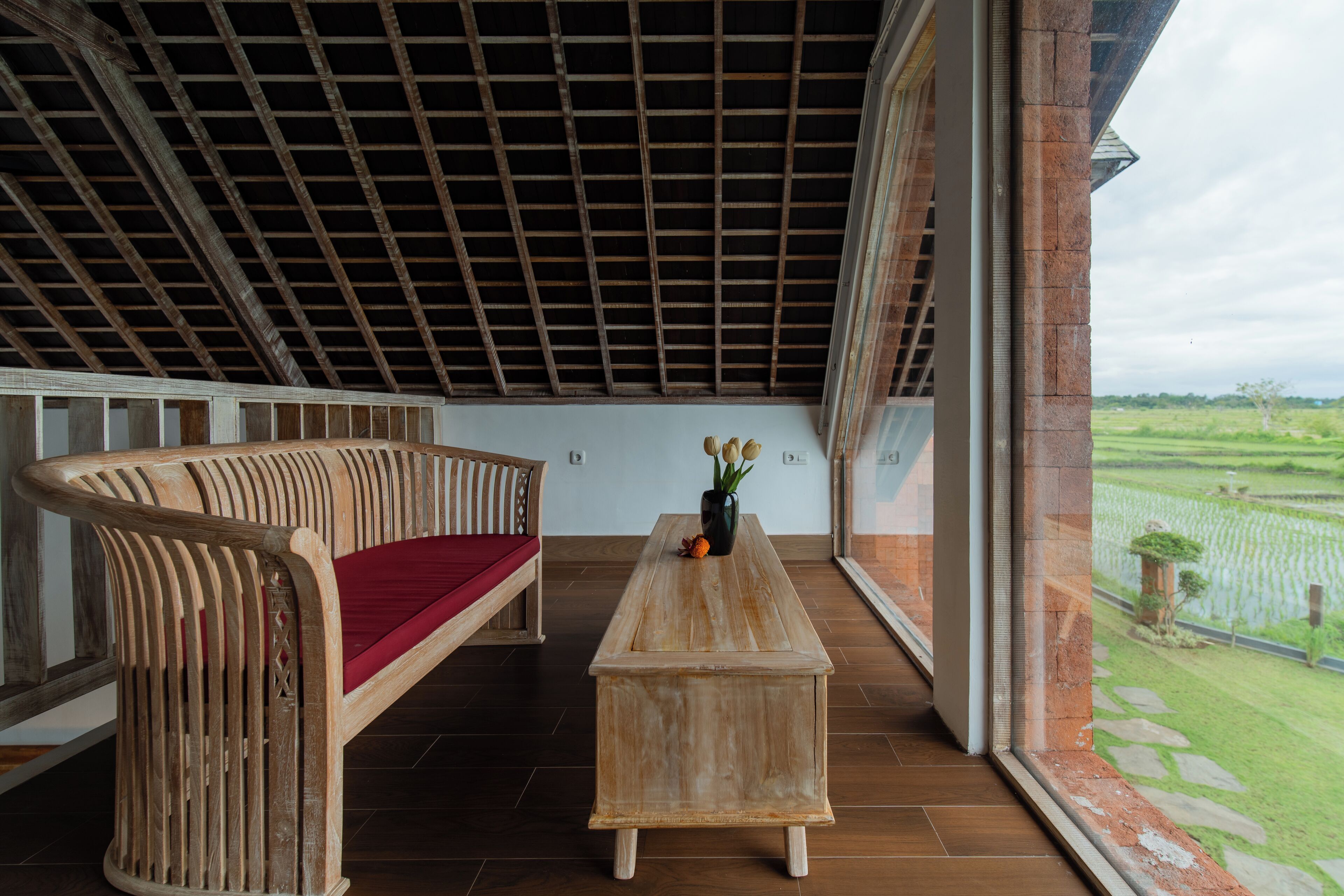 A wooden bench with a red cushion, a wooden table with a vase of flowers, and a view of a green landscape through large windows.