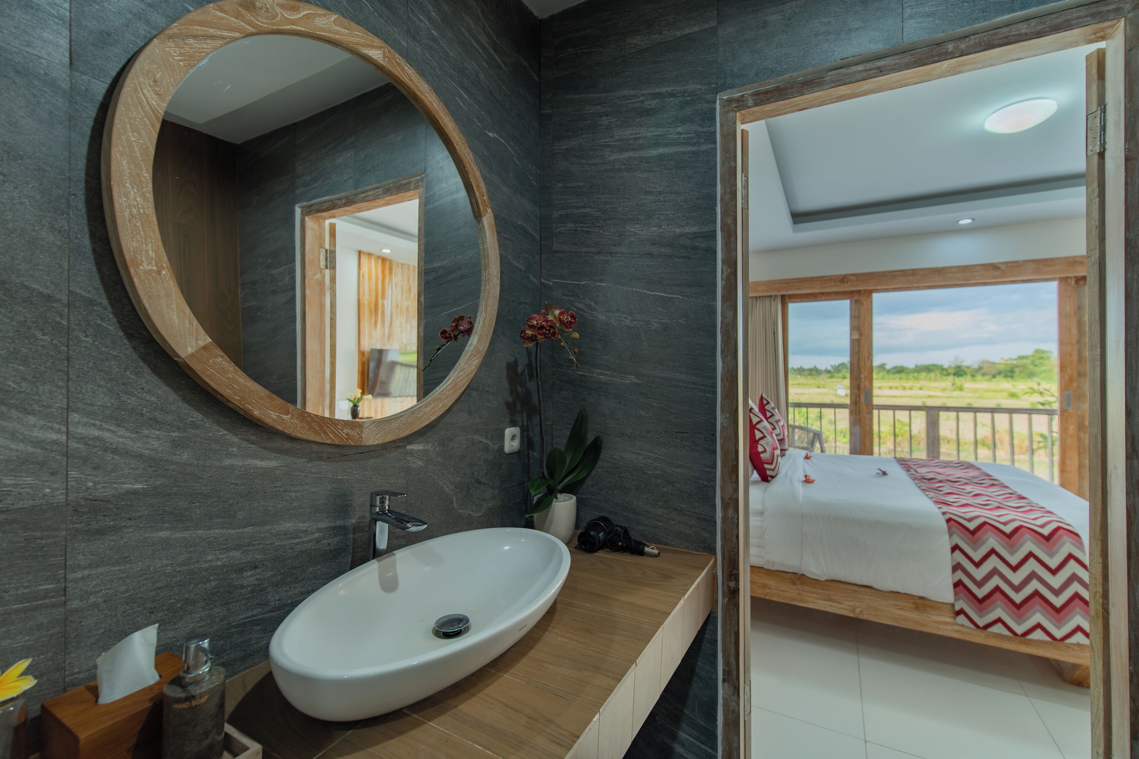 A modern bathroom with a large oval sink, a wooden-framed mirror, and a view of a balcony with a bed.