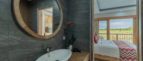 A modern bathroom with a large oval sink, a wooden-framed mirror, and a view of a balcony with a bed.
