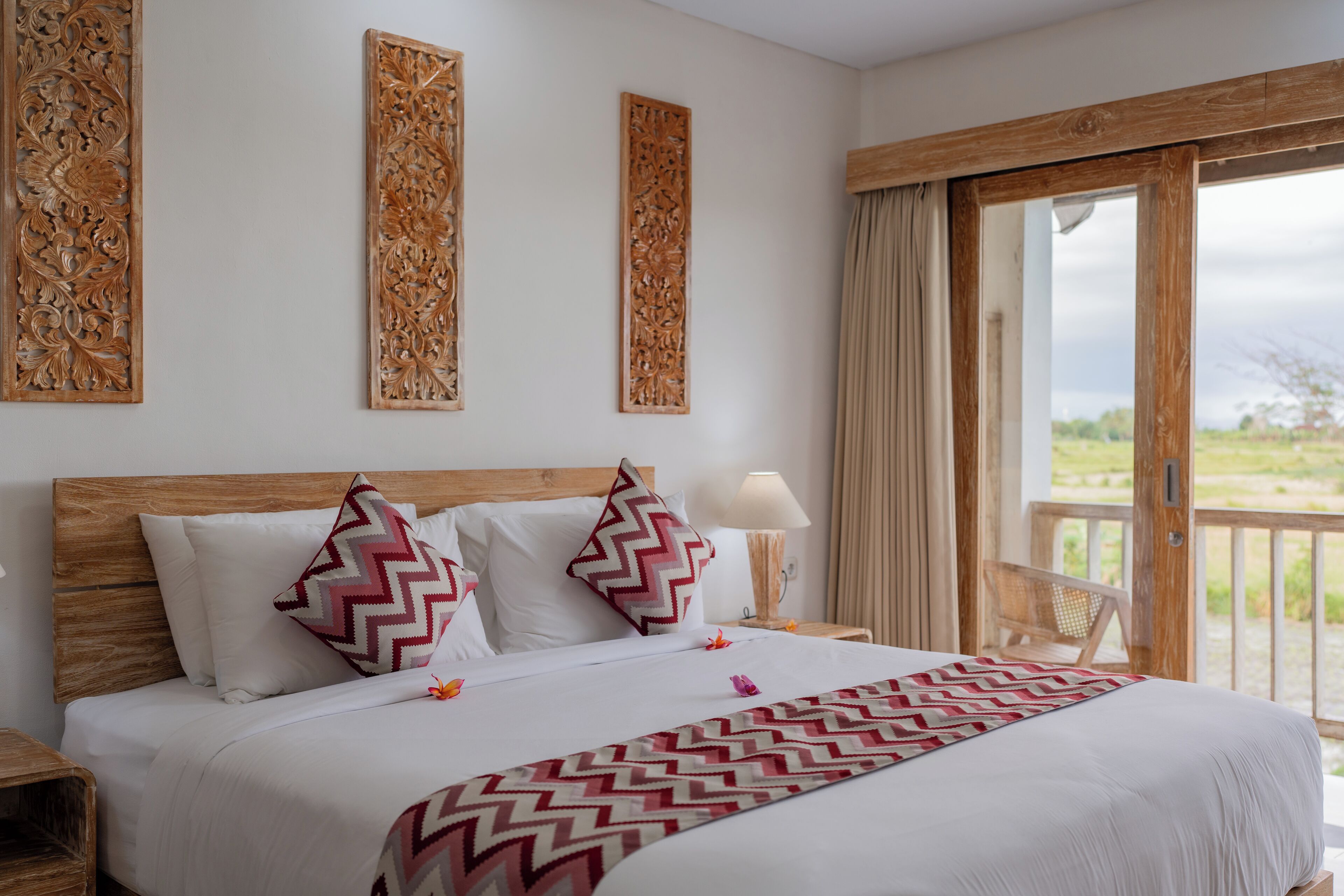 A hotel room with a wooden headboard, a large bed with white bedding, decorative wall panels, a nightstand with a lamp, and a view of the outdoors through a sliding glass door.