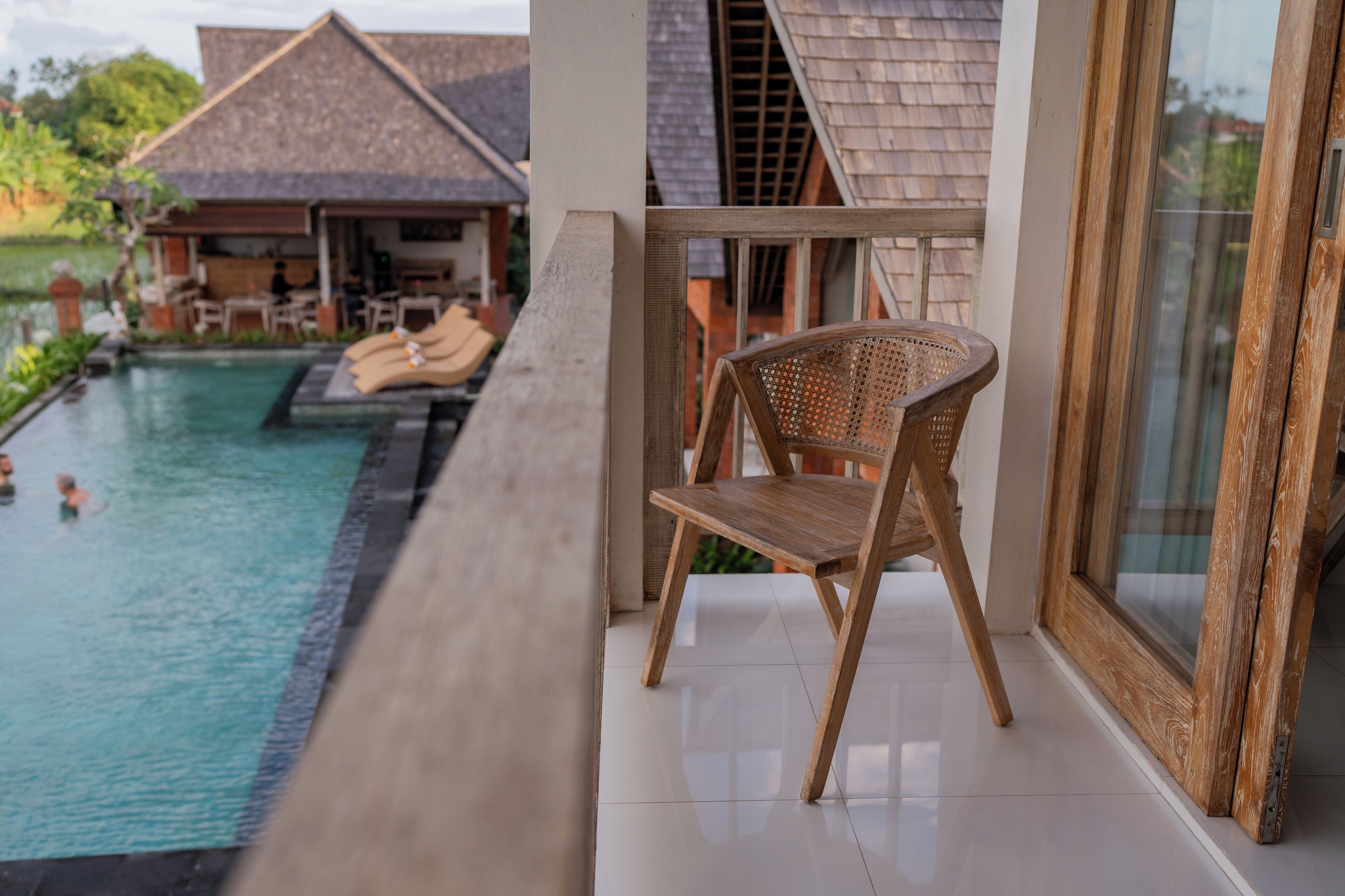 Une chaise en bois près d’une piscine, une porte vitrée et, au fond, un bâtiment avec un toit en tuiles.
