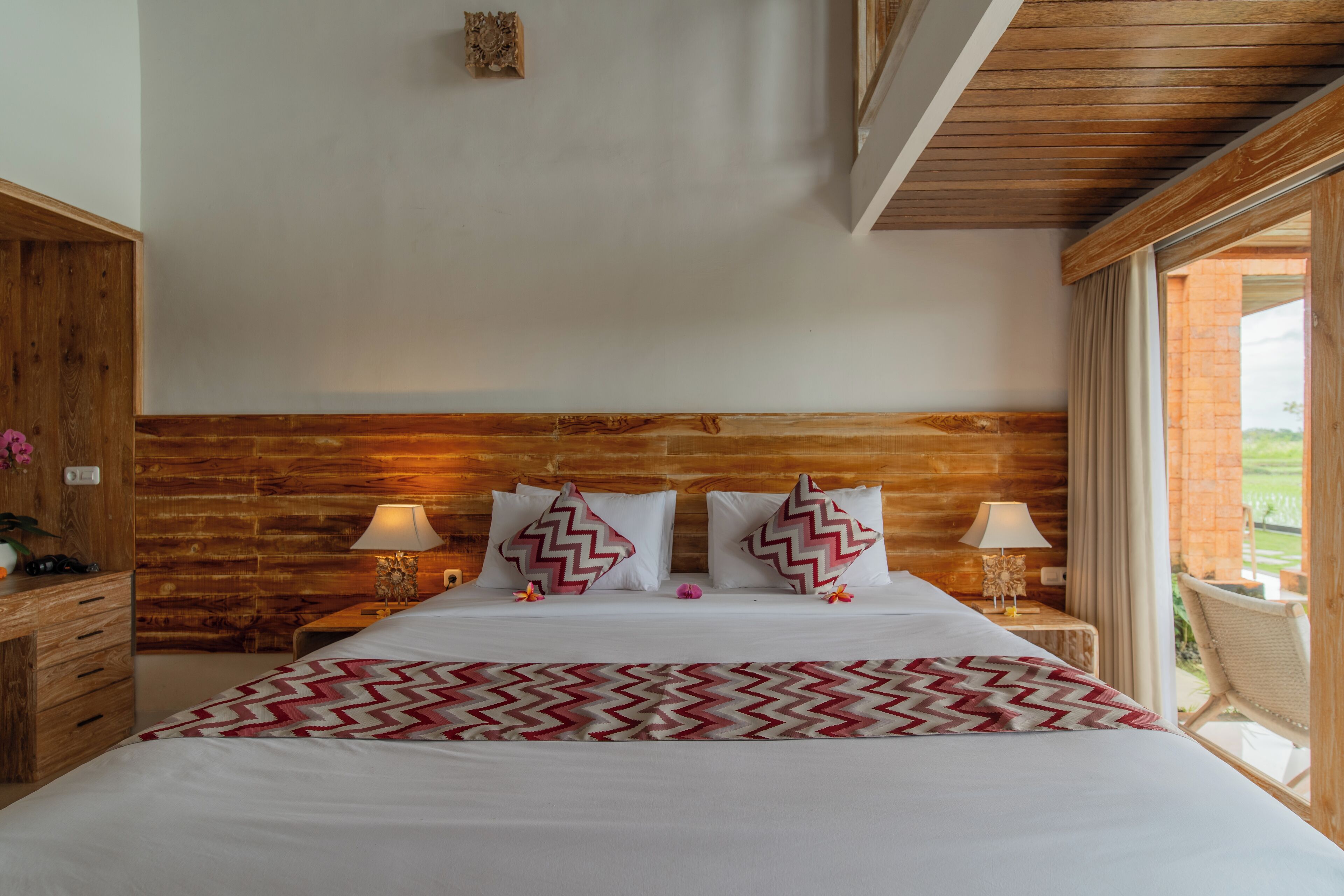 A bedroom with a wooden headboard, two bedside tables with lamps, a bed with patterned pillows, and a view of the outdoors.