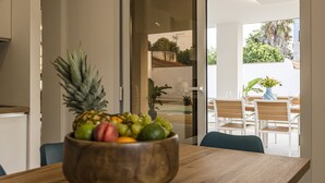 Une table à manger en bois, avec un saladier de fruits placé au centre, une porte vitrée donnant sur un espace extérieur aménagé avec des chaises et une table, et une vue sur la verdure environnante.