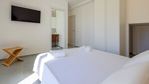 Une chambre d’hôtel moderne avec un grand lit, un téléviseur à écran plat fixé au mur, une table de chevet en bois et une salle de bain visible à travers une porte ouverte.