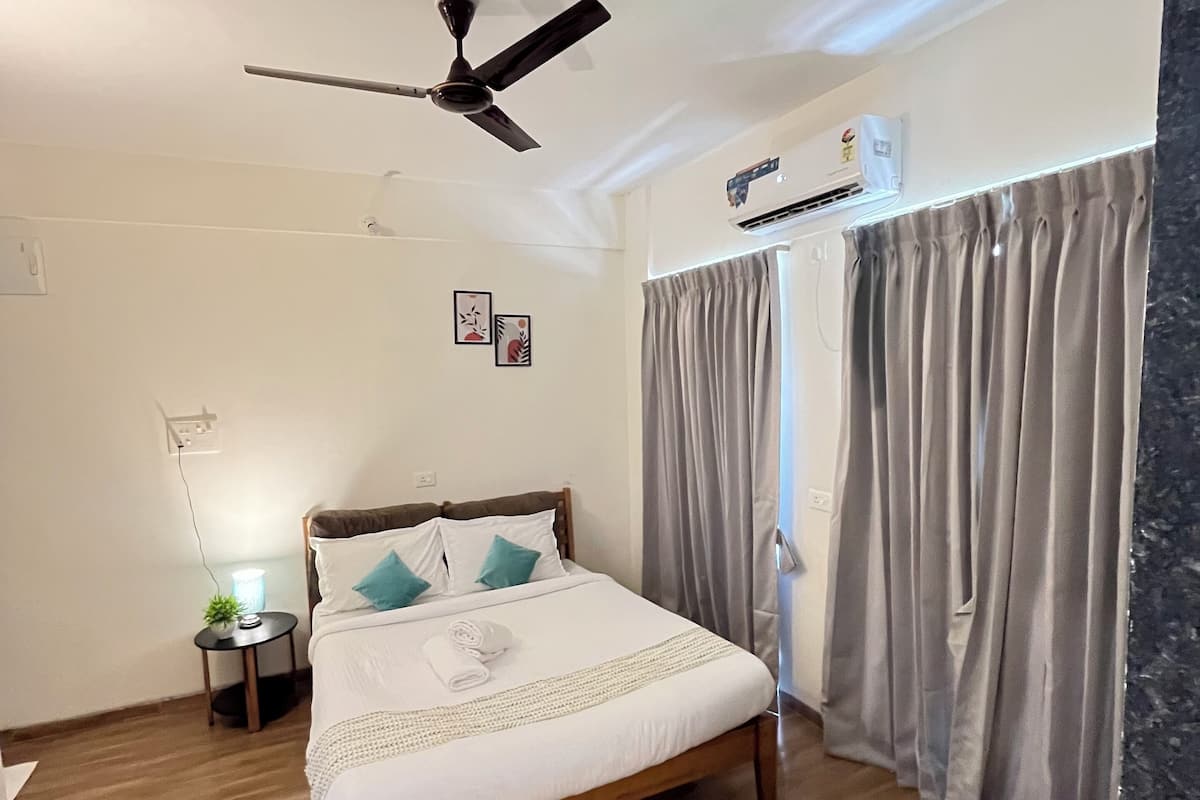 A bedroom with a bed, a ceiling fan, an air conditioning unit, a nightstand with a lamp, and a window with curtains.