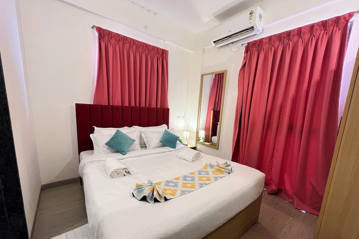 A hotel room with a large bed, red curtains, a white headboard, and an air conditioning unit.