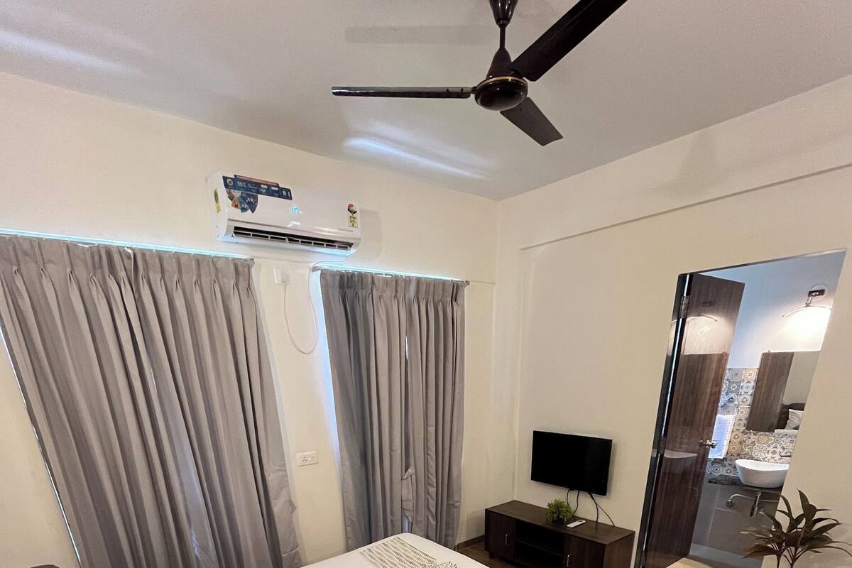 A modern bedroom with a bed, curtains, a ceiling fan, an air conditioning unit, a TV, a small table with a lamp, and a potted plant.