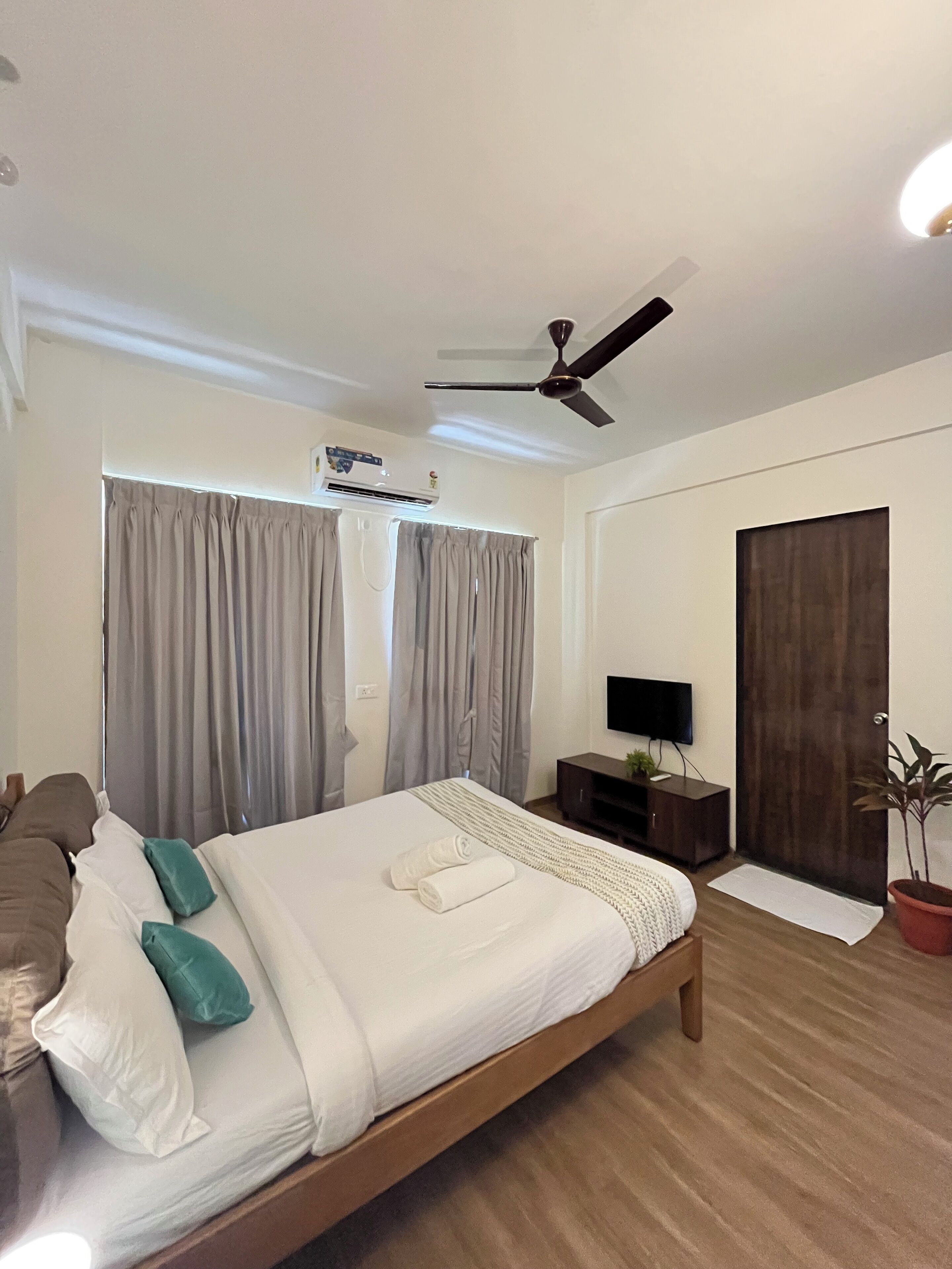 Ein modernes Hotelzimmer mit einem Bett, einem Fernseher, einem Deckenventilator, einer Klimaanlage, einem dunklen Holzschrank und einer Topfpflanze.