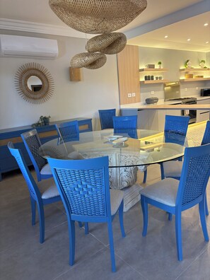 Un espace repas moderne avec une table en verre entourée de chaises bleues, un luminaire suspendu en fibres tressées et une cuisine en arrière-plan.