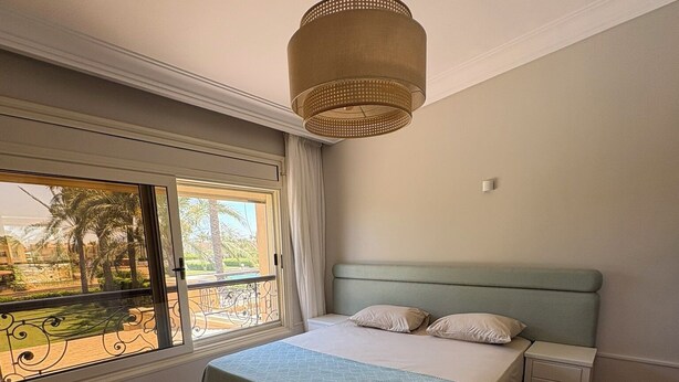 A bedroom with a bed, a nightstand, a window with a view of palm trees, and a hanging light fixture.