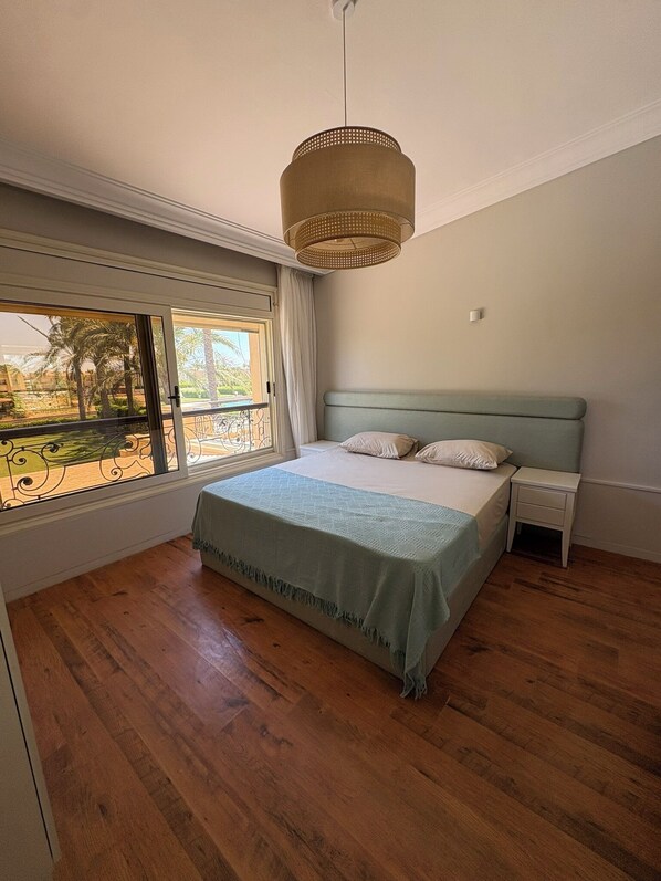 A bedroom with a bed, a nightstand, a window with a view of palm trees, and a hanging light fixture.
