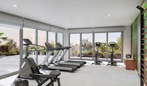 Un gimnasio moderno con cintas de correr, bicicletas estáticas y pesas libres. Amplias ventanas ofrecen vistas a zonas verdes.