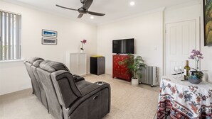 A living room with a reclining sofa, a ceiling fan, a flat-screen TV, a red cabinet, a potted plant, and a table with a floral tablecloth.