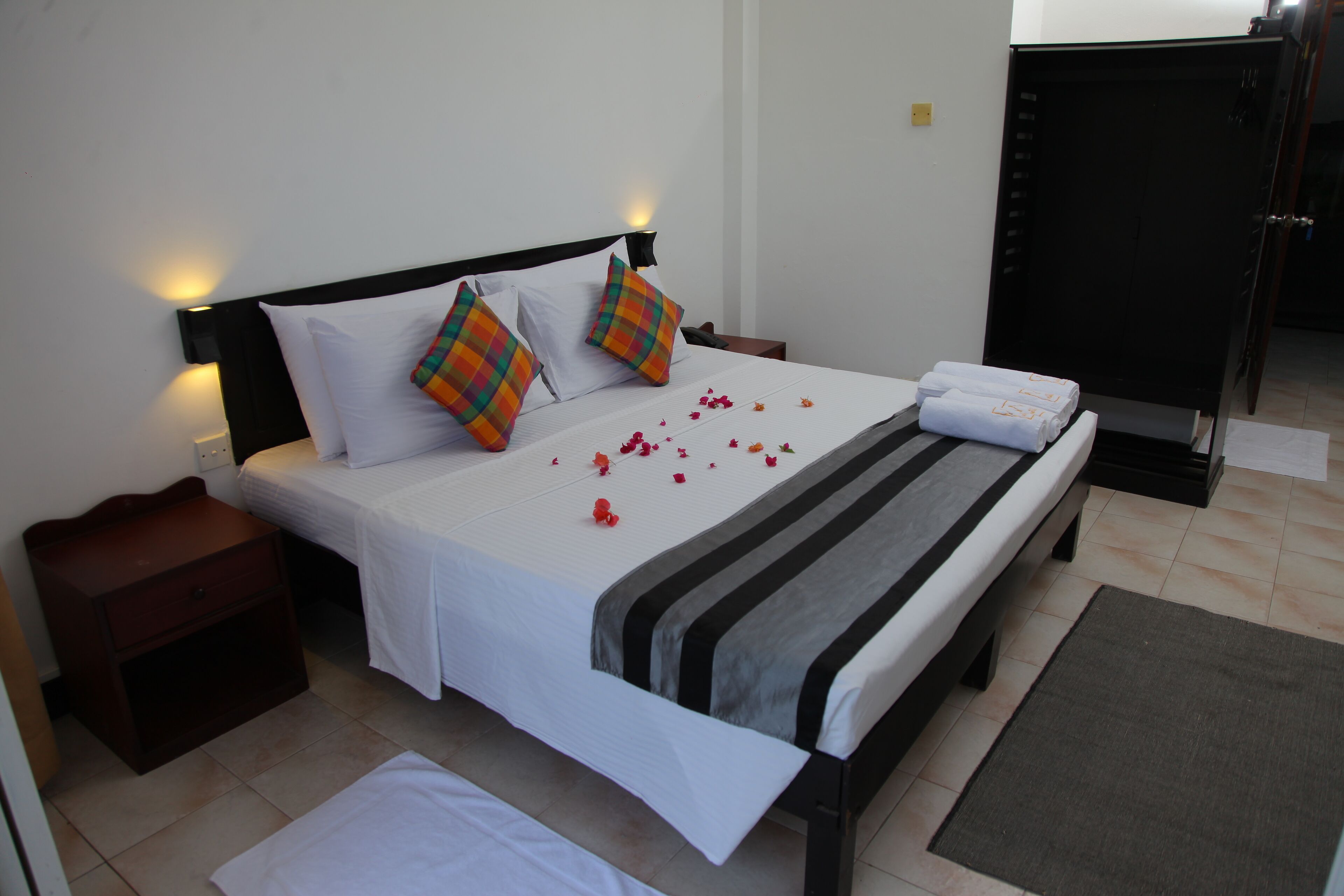 A neatly made bed with white linens, two colorful pillows, and a striped blanket. There are rose petals scattered on the bed and a small nightstand with a drawer.