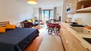 A compact hotel room with a bed, a dining table, chairs, a kitchen area with a sink and cabinets, and a large window with curtains. - Bad Gastein Fallside Apartment (Bad Kleinkirchheim)