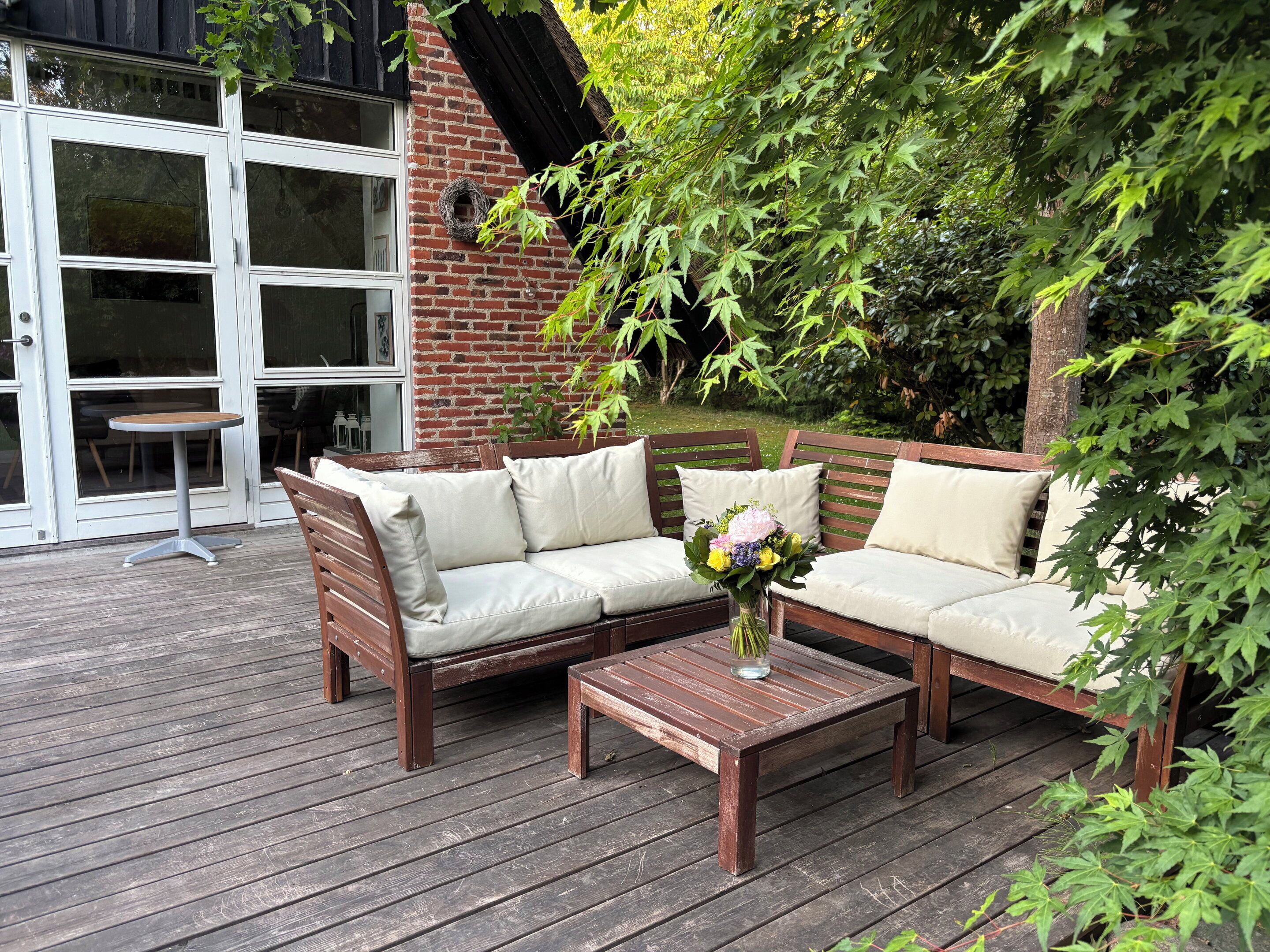 Um deck de madeira com um sofá e uma mesa, rodeado por vegetação.