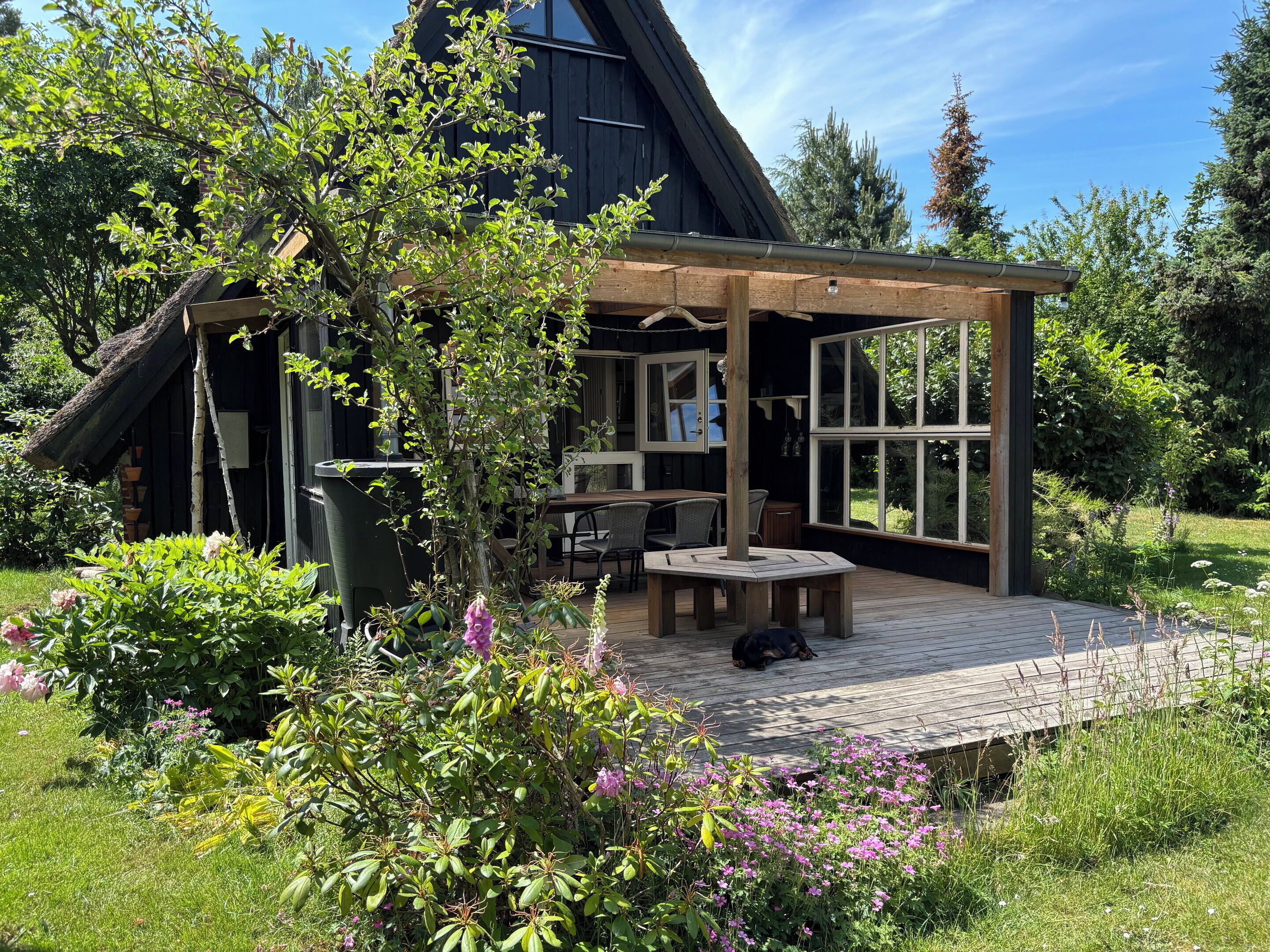 En træterrasse med et bord og stole, omgivet af en have med forskellige planter og blomster.