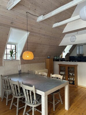 A dining area with a high ceiling, wooden beams, a table with chairs, a hanging light fixture, and a view through a window.