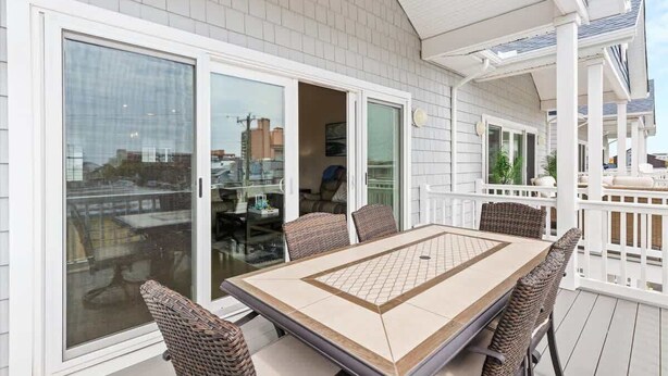 Um pátio externo coberto com uma mesa de jantar e cadeiras, grandes portas de vidro deslizantes e um guarda-corrimão branco.