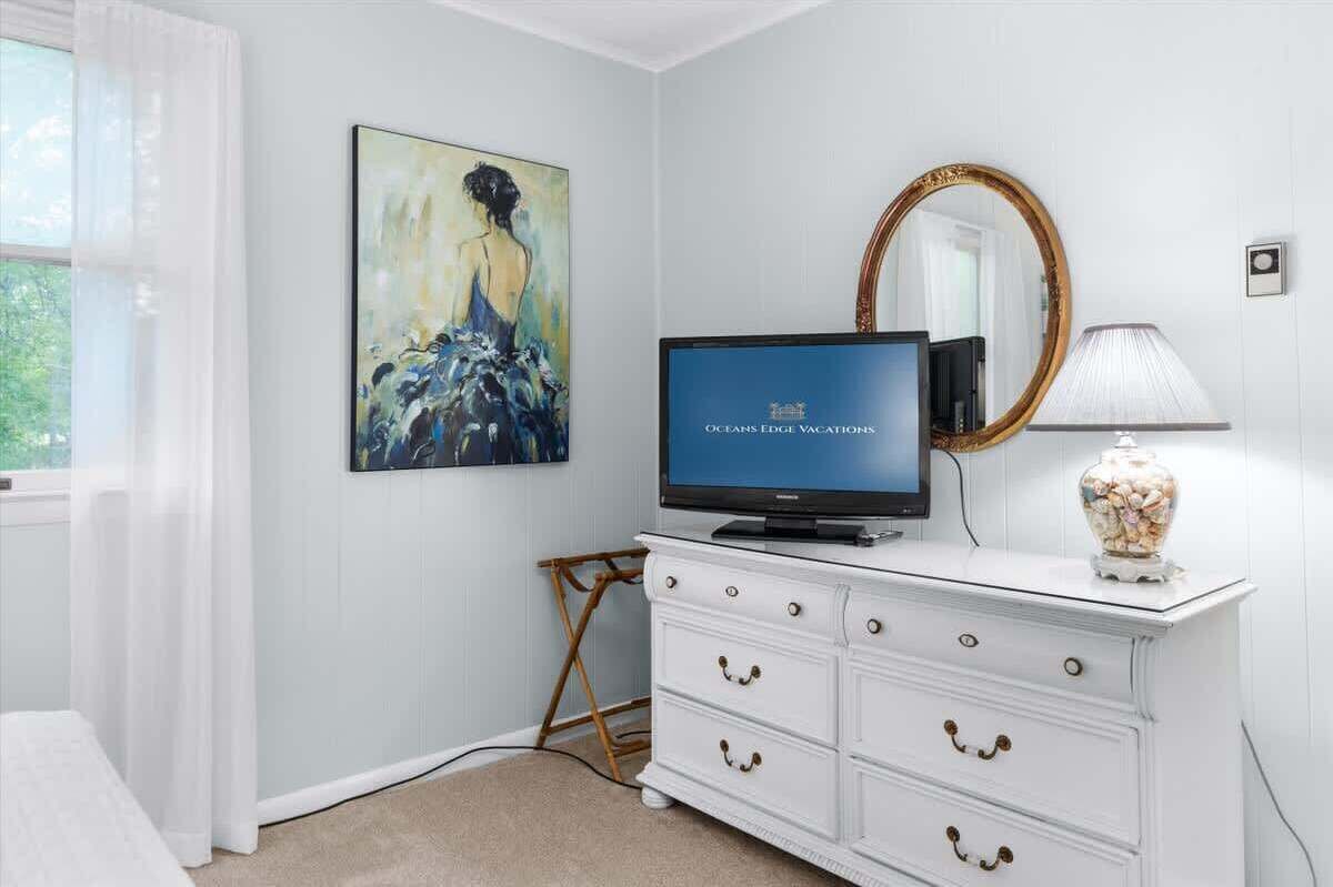 A bedroom with a television, a white dresser, a painting, and a mirror.