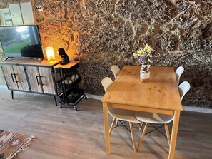 A wooden dining table with chairs, a television on a cabinet, and a stone wall.