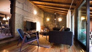 A cozy living room with a stone wall, wooden ceiling beams, a fireplace, and a large window.