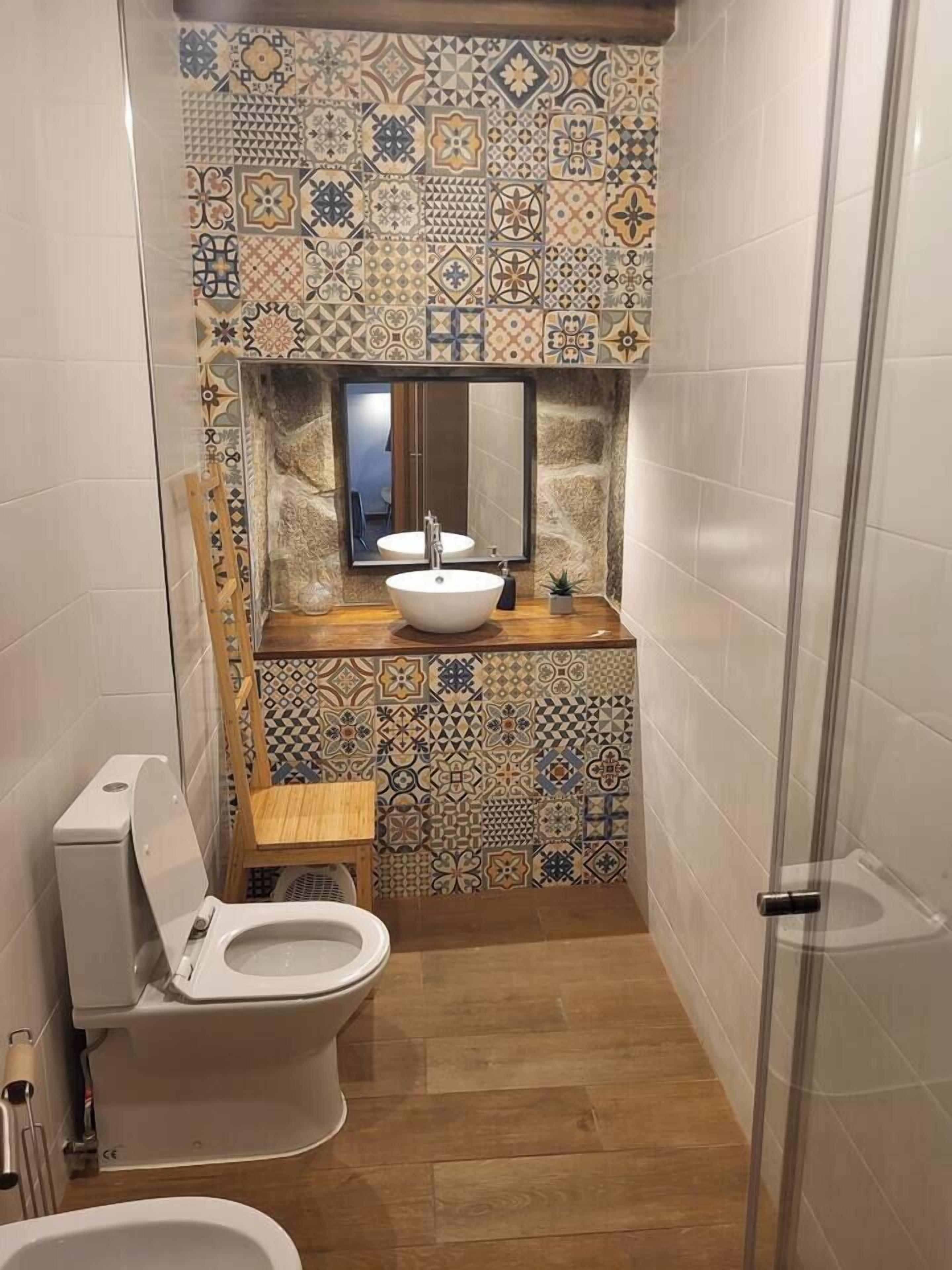A small bathroom with a toilet, sink, and shower. The walls are tiled with a geometric pattern.