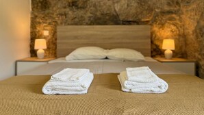 Una cama bien hecha con dos toallas blancas dobladas sobre una superficie texturizada, flanqueada por dos lámparas de noche en mesitas a juego, con un fondo de pared de piedra.