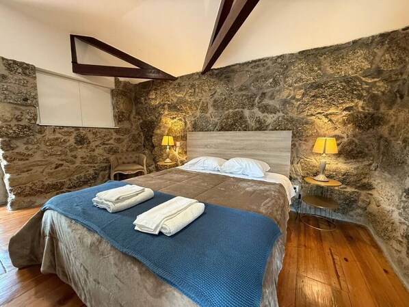 Une chambre avec un mur en pierre, un lit recouvert d’une couverture bleue, deux lampes de chevet et un plancher en bois.