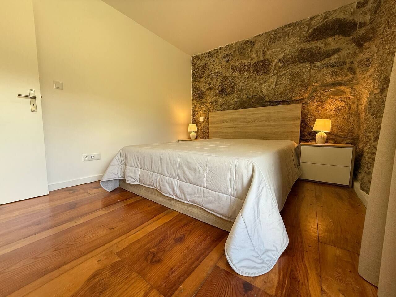 Un dormitorio con suelo de madera, una cama con colcha blanca, una pared de piedra y una mesita de noche con lámpara.