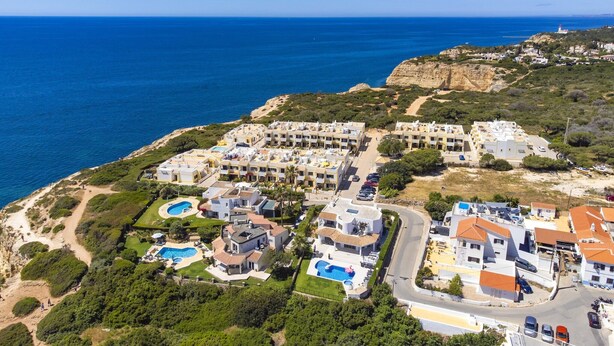 An aerial view of a coastal residential area with multiple apartment buildings, a swimming pool, and a lighthouse in the background. - Apartment with beautiful terrace, sea-views (Benagil)