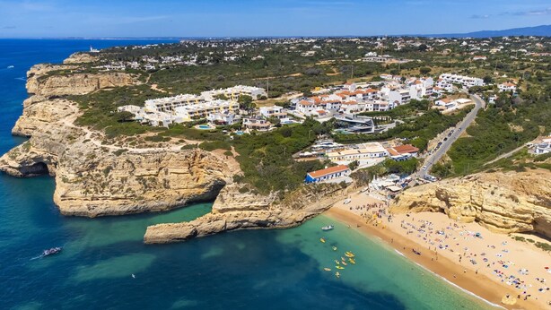 An aerial view of a coastal town with buildings clustered on a cliff, a beach with people and boats, and clear blue waters. - Charming apartment near Atlantic Ocean (Benagil)
