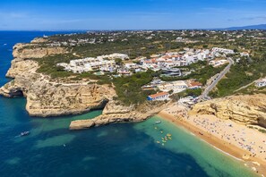 An aerial view of a coastal town with buildings clustered on a cliff, a beach with people and boats, and clear blue waters.