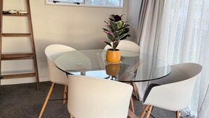 A modern dining area with a glass table, white chairs, a potted plant, a ladder, and a window with curtains. - Palm Haven Beach Retreat (Paraparaumu Beach)