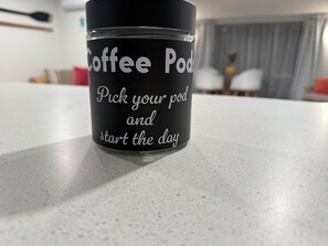 A coffee pod container with the text "Coffee Pod Pick your pod and start the day" on a countertop. - Palm Haven Beach Retreat (Paraparaumu Beach)