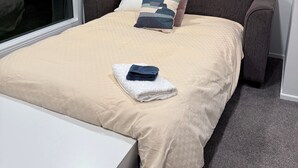 A neatly made bed with a beige bedspread, two pillows, and a folded towel. - Palm Haven Beach Retreat (Paraparaumu Beach)