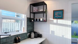 A kitchen with a window, a kettle, a coffee maker, and a shelf with a box and a plant. - Palm Haven Beach Retreat (Paraparaumu Beach)