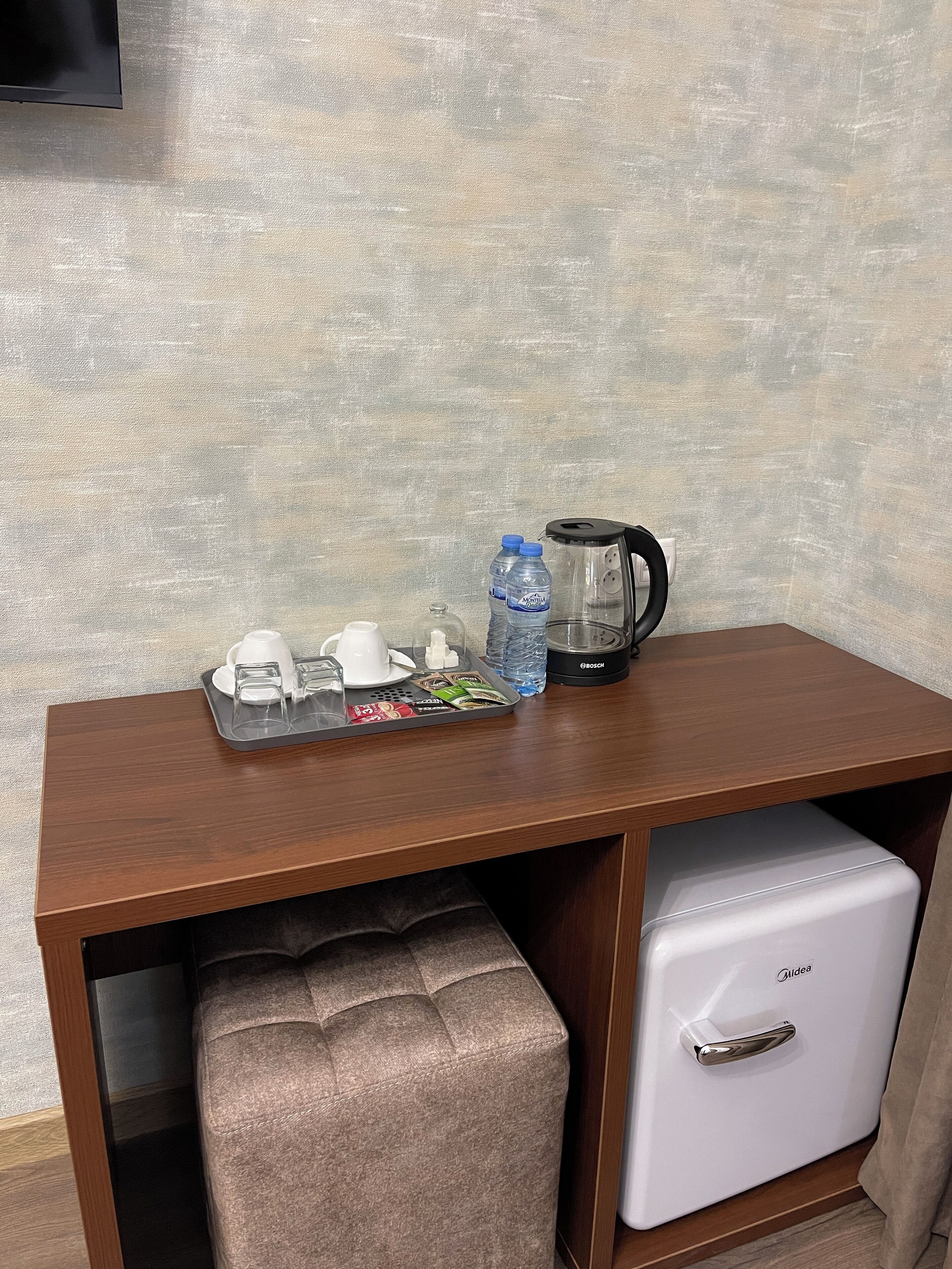 Un escritorio de madera con una bandeja que contiene una cafetera, botellas de agua y una taza. Abajo del escritorio, hay un refrigerador blanco.